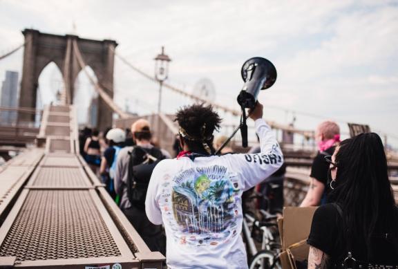 Aktivist mit einem Megaphon in der ausgestreckten rechten Hand, der in einer Gruppe Menschen über die Brooklyn Bridge läuft.
